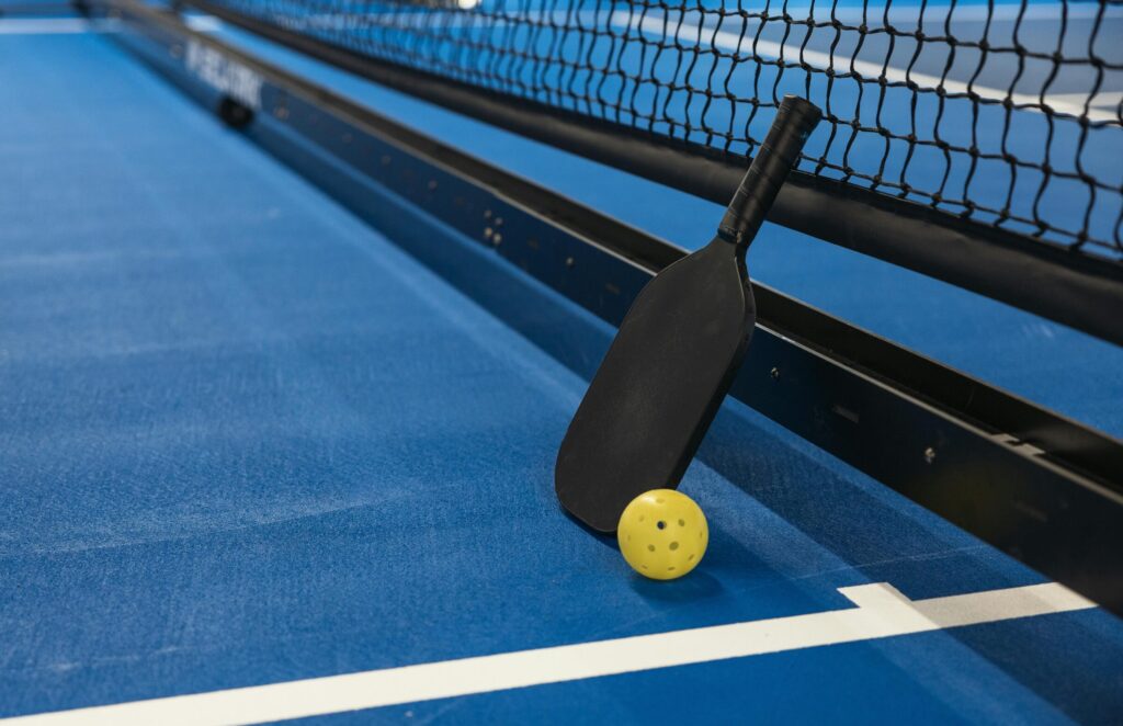 Pickleball paddle resting against net with yellow ball on blue court