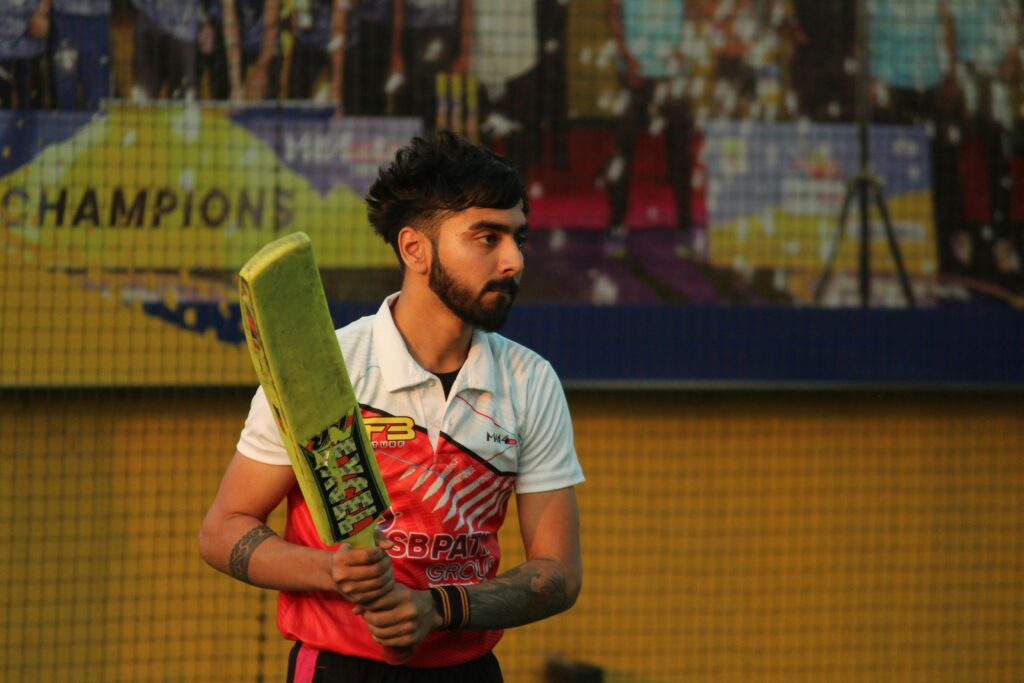 A focused cricketer holding a bat, ready to play on an artificial turf field. The backdrop includes a colorful advertisement and a netted area.